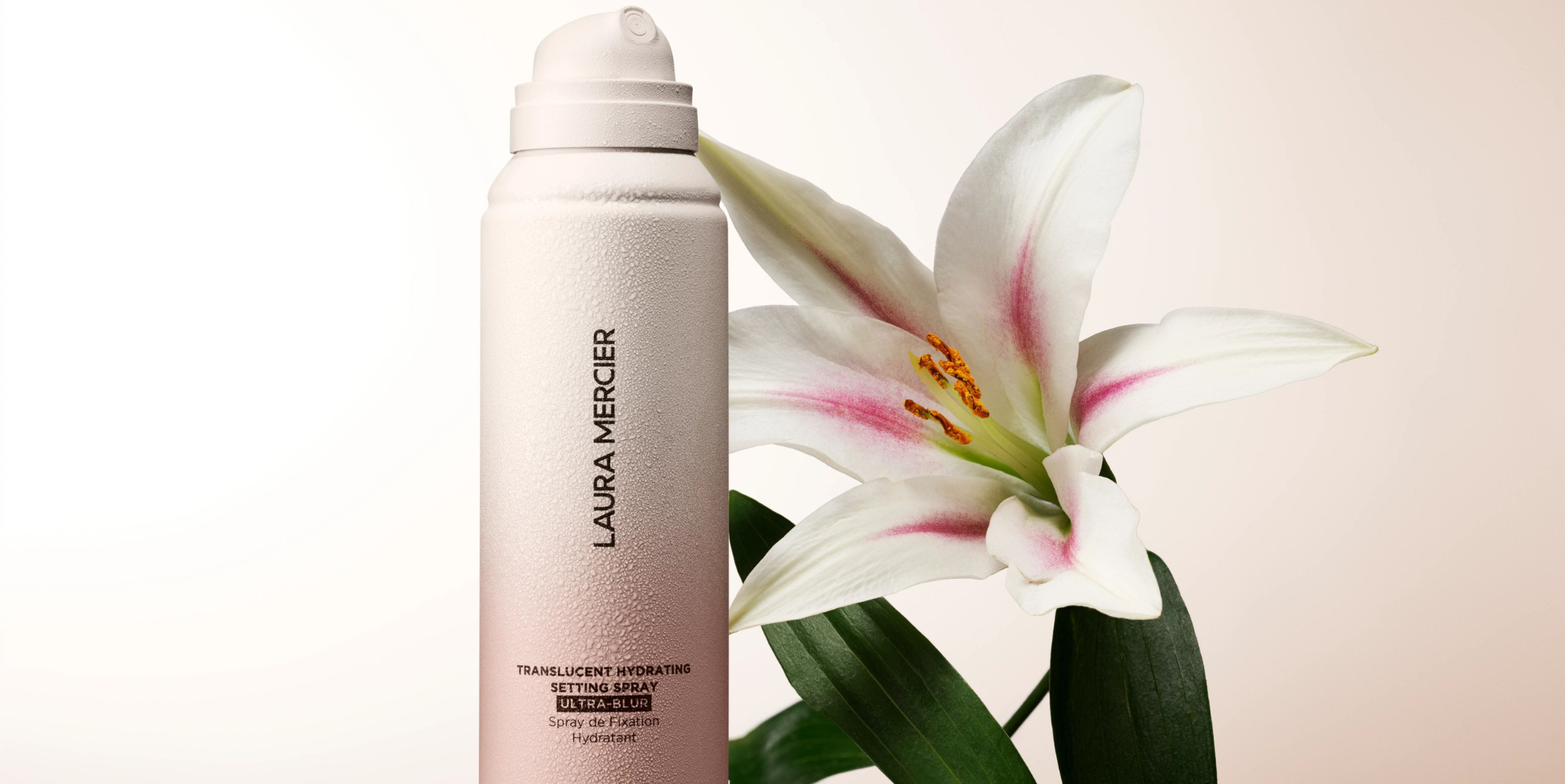 Laura Mercier's Translucent hydrating Setting Spray next to a White Lily on a gradient light beige background.