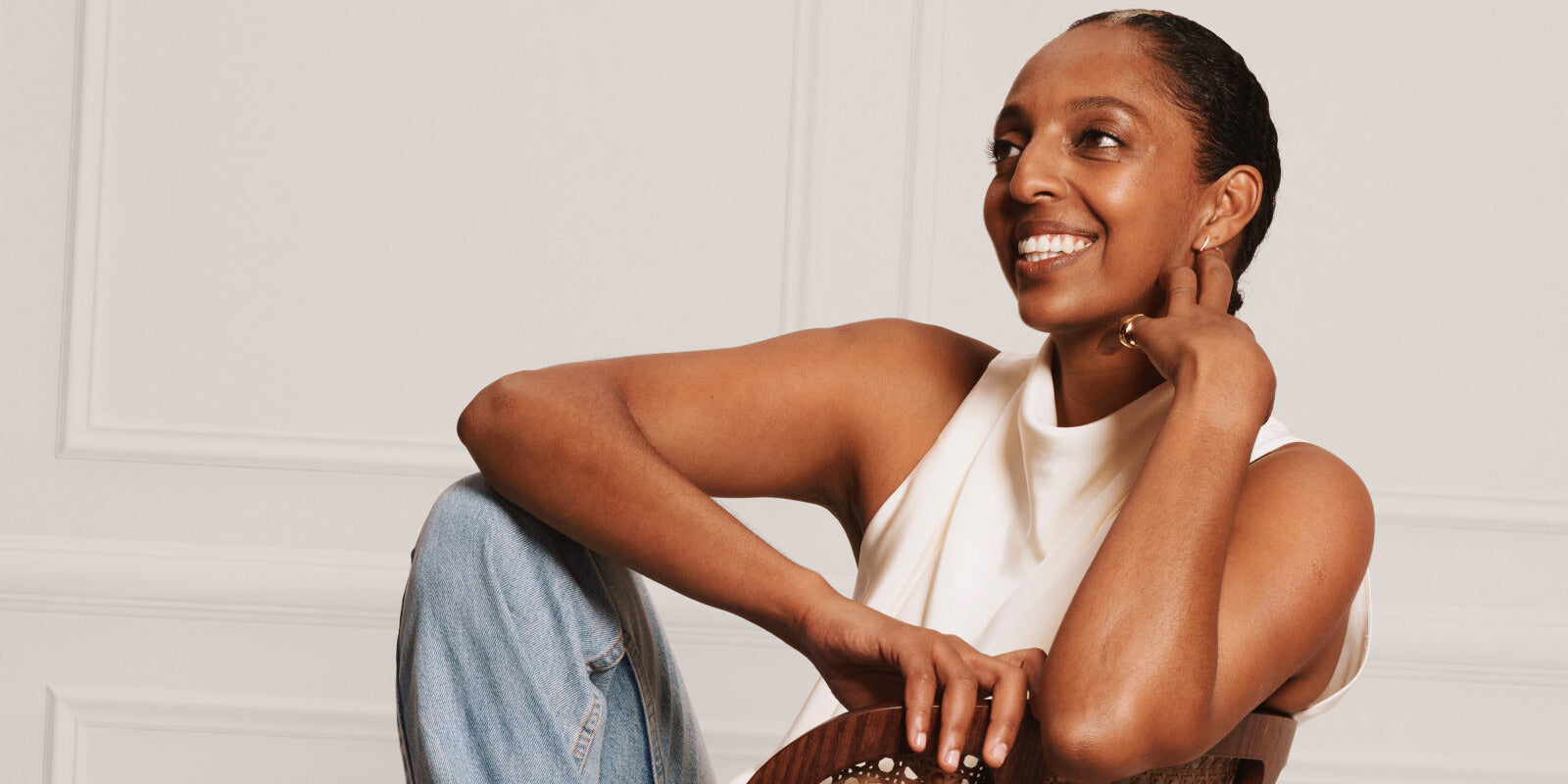 Dark-skinned model with white halter top and jeans sitting on a chair smiling