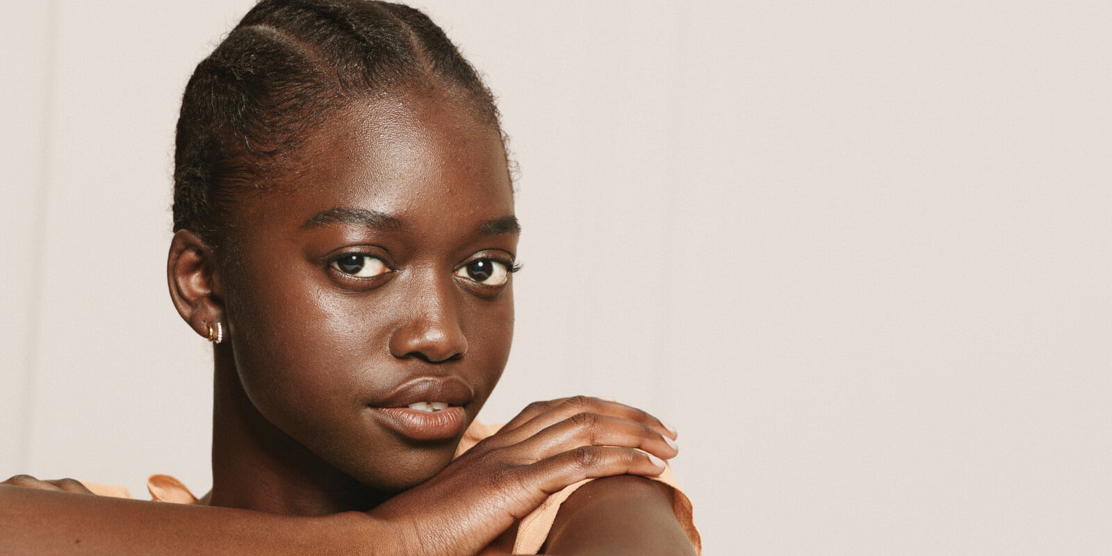 Dark skinned model in front of beige background with her hand on her shoulder