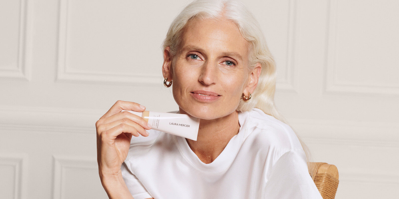 Pale middle-aged model with white hair in a white T-shirt holding up Laura Mercier tinted moisturizer